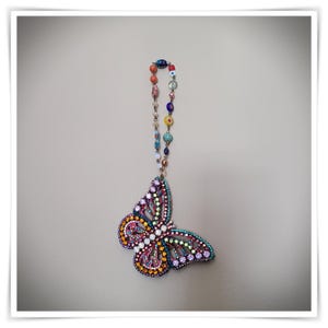 May include: A colorful butterfly pendant with a beaded chain. The butterfly is adorned with a variety of gemstones in shades of pink, purple, yellow, and turquoise. The beaded chain features a mix of colorful beads.