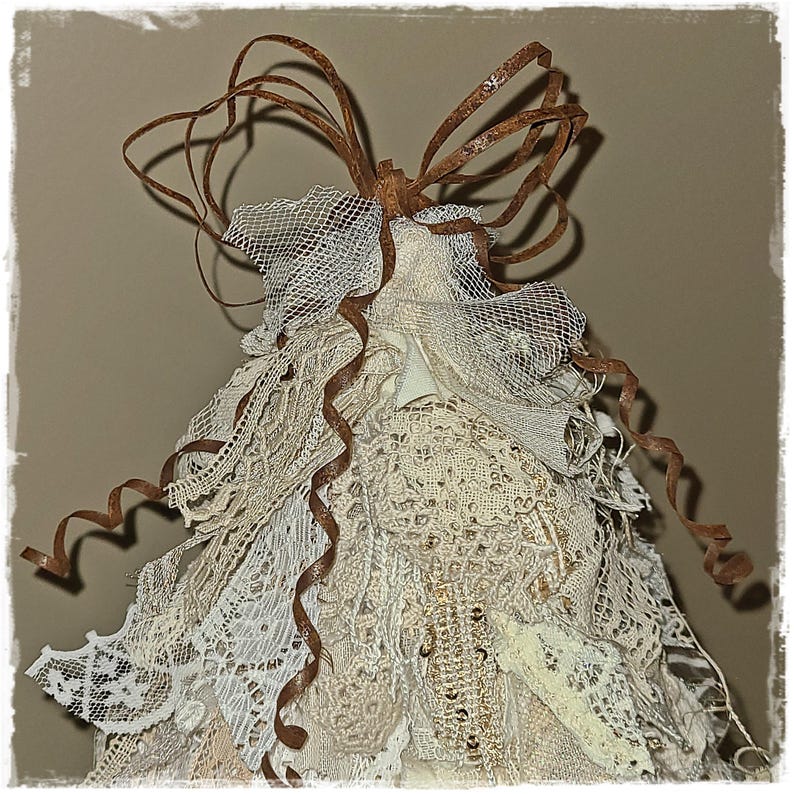 May include: A handmade Christmas tree ornament. The tree is made of layers of cream-colored lace and netting, with brown ribbon accents and a brown, curly bow at the top. The ornament has a vintage, rustic aesthetic.