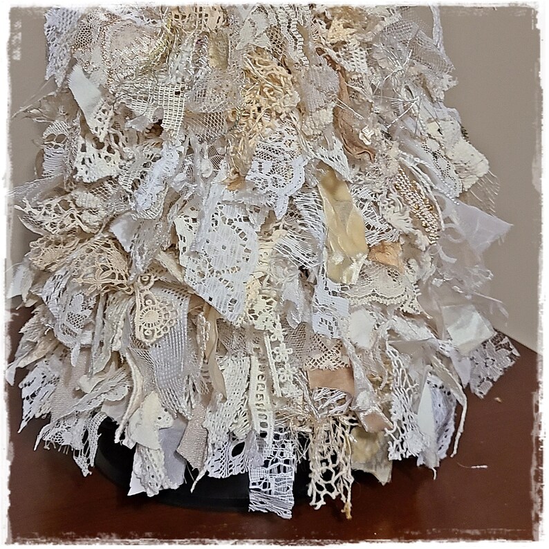 May include: A handcrafted, layered textile art piece. The artwork is composed of various lace and fabric scraps in shades of white, cream, and beige. The piece has a textured, three-dimensional appearance, with a variety of lace patterns and textures.