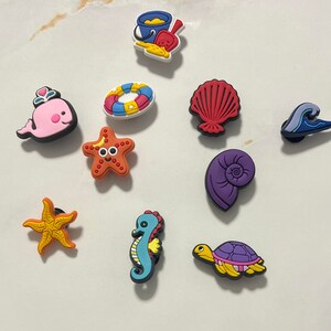 May include: A collection of colorful beach-themed charms, including a pink whale, starfish, seahorse, and a turtle. Other charms include a seashell, wave, life preserver, sand pail, and a spiral shell. These charms are perfect for decorating shoes or accessories.
