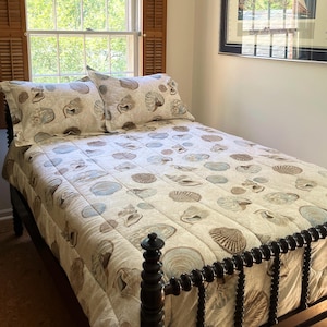 May include: A bed featuring a dark brown wooden spindle frame and a quilt set with two matching pillows. The bedding is cream-colored and decorated with a repeating pattern of various seashells in shades of brown, tan, and soft blue throughout.