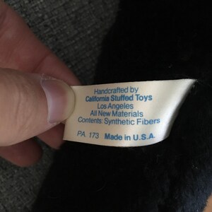 Op de afbeelding: Een close-up van een wit label met blauwe tekst, vastgehouden door een hand, tegen een zwarte pluche achtergrond. Het label luidt "Handcrafted by California Stuffed Toys, Los Angeles, All New Materials, Contents: Synthetic Fibers, PA 173 Made in U.S.A."