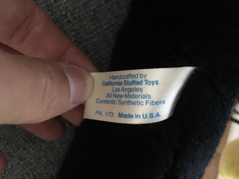 Op de afbeelding: Een close-up van een wit label met blauwe tekst, vastgehouden door een hand, tegen een zwarte pluche achtergrond. Het label luidt "Handcrafted by California Stuffed Toys, Los Angeles, All New Materials, Contents: Synthetic Fibers, PA 173 Made in U.S.A."