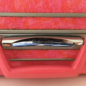Pu&ograve; includere: Una valigia rosa brillante dalla superficie testurizzata, caratterizzata da un bordo in metallo argentato e una maniglia in plastica rosa coordinata. Il marchio Samsonite &egrave; impresso in rilievo sulla parte argentata sopra la maniglia.