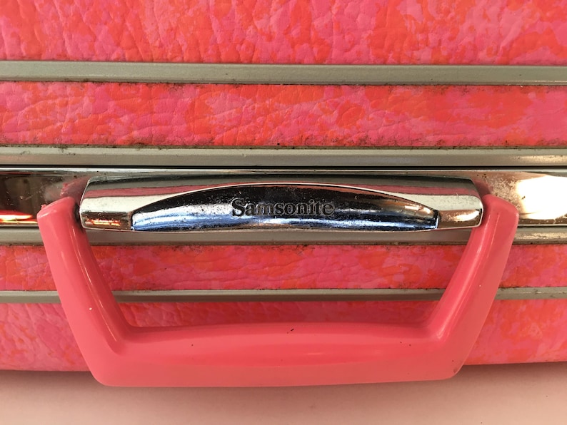 Pu&ograve; includere: Una valigia rosa brillante dalla superficie testurizzata, caratterizzata da un bordo in metallo argentato e una maniglia in plastica rosa coordinata. Il marchio Samsonite &egrave; impresso in rilievo sulla parte argentata sopra la maniglia.