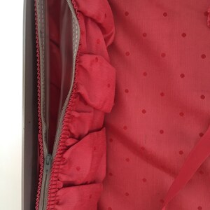 Pu&ograve; includere: L'interno di una valigia caratterizzato da una fodera in tessuto rosso vivace con un sottile motivo a pois tono su tono. Uno scomparto laterale &egrave; chiuso da una cerniera e presenta un bordo in tessuto arricciato.