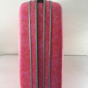 Pu&ograve; includere: Una valigia rigida rettangolare caratterizzata da una vivace texture screziata rosa e arancione. L'esterno &egrave; diviso da due strisce decorative verticali grigio metallizzato al centro, con un piccolo meccanismo di chiusura visibile in alto.