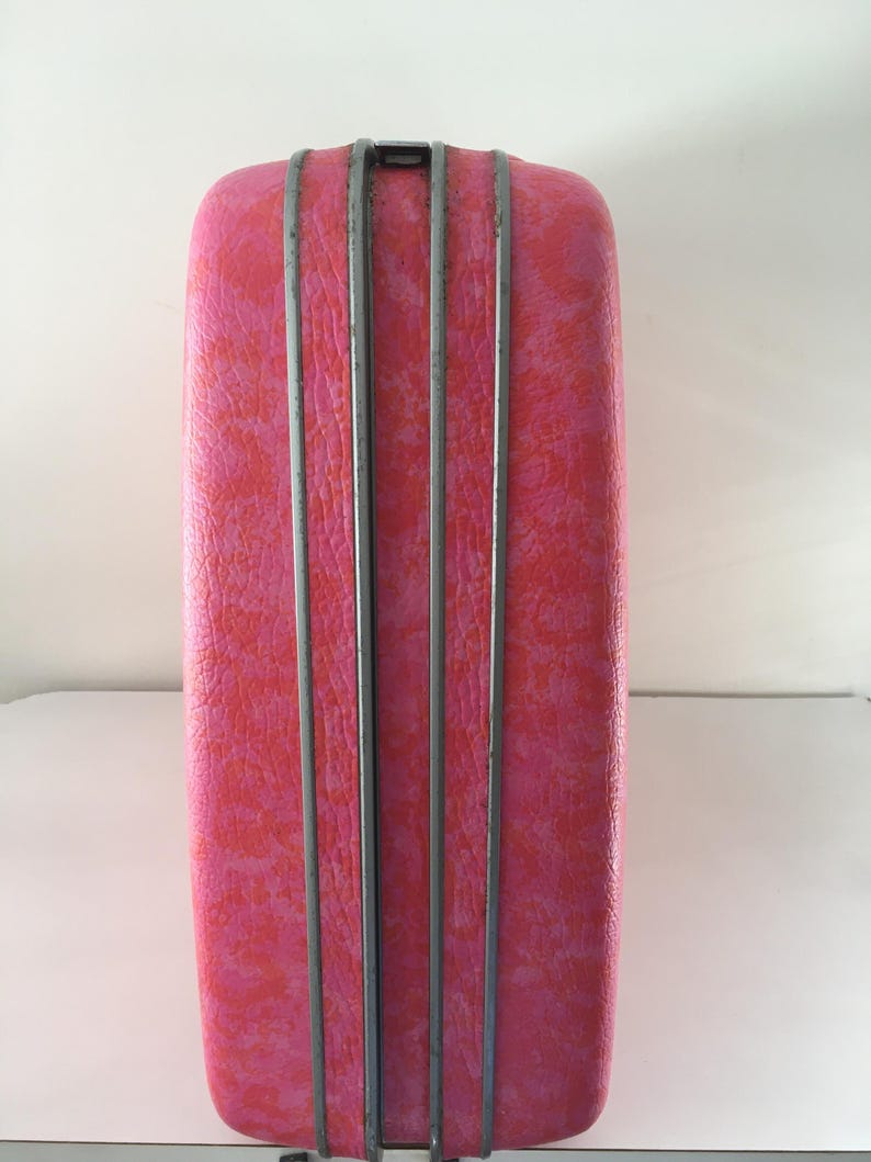 Pu&ograve; includere: Una valigia rigida rettangolare caratterizzata da una vivace texture screziata rosa e arancione. L'esterno &egrave; diviso da due strisce decorative verticali grigio metallizzato al centro, con un piccolo meccanismo di chiusura visibile in alto.