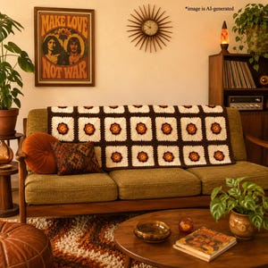 May include: A rectangular crochet throw blanket featuring a repeating pattern of cream-colored squares with dark brown borders and a central orange and yellow flower motif, draped over a textured olive green sofa with two decorative throw pillows.