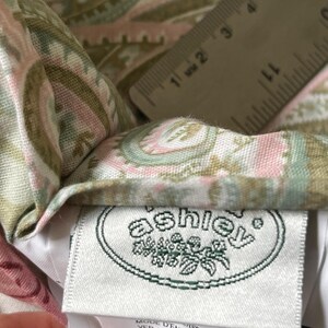 May include: A fabric textile featuring a paisley pattern in shades of sage green, soft pink, and cream. A white clothing label with the text 'ashley' in a green oval logo is attached. A ruler shows a measurement of approximately 1.5 inches.