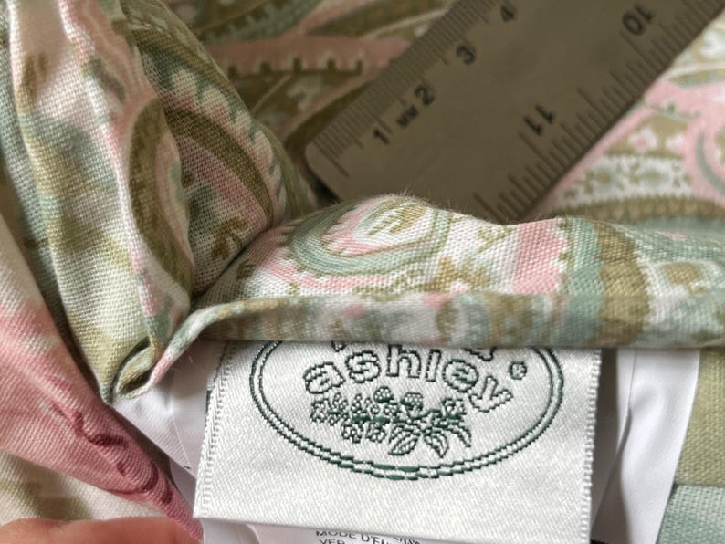 May include: A fabric textile featuring a paisley pattern in shades of sage green, soft pink, and cream. A white clothing label with the text 'ashley' in a green oval logo is attached. A ruler shows a measurement of approximately 1.5 inches.