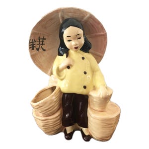 May include: A ceramic figurine of a woman in a yellow jacket and dark pants, holding an umbrella. The figurine has a stack of baskets and jars. The umbrella has Chinese characters on it.
