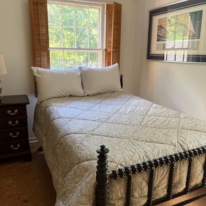 May include: A bedroom scene featuring a dark wood spindle bed frame with a light green and white paisley quilted bedspread and two white pillows. A dark wood four-drawer nightstand holds a blue and white lamp with a beige fabric shade.
