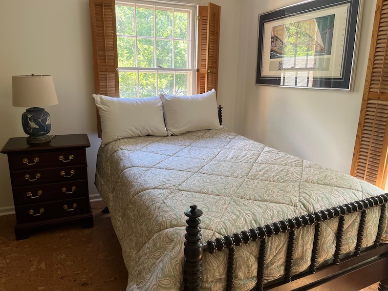 May include: A bedroom scene featuring a dark wood spindle bed frame with a light green and white paisley quilted bedspread and two white pillows. A dark wood four-drawer nightstand holds a blue and white lamp with a beige fabric shade.