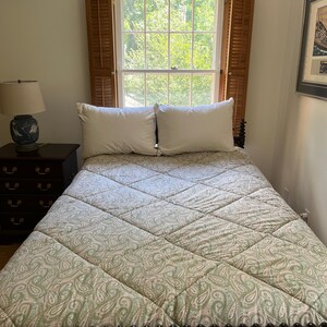 May include: A bed featuring a quilted comforter with a light green and white paisley pattern, two white pillows, and a dark wood nightstand with a blue and white ceramic lamp. The bed is positioned in front of a window with wooden shutters.