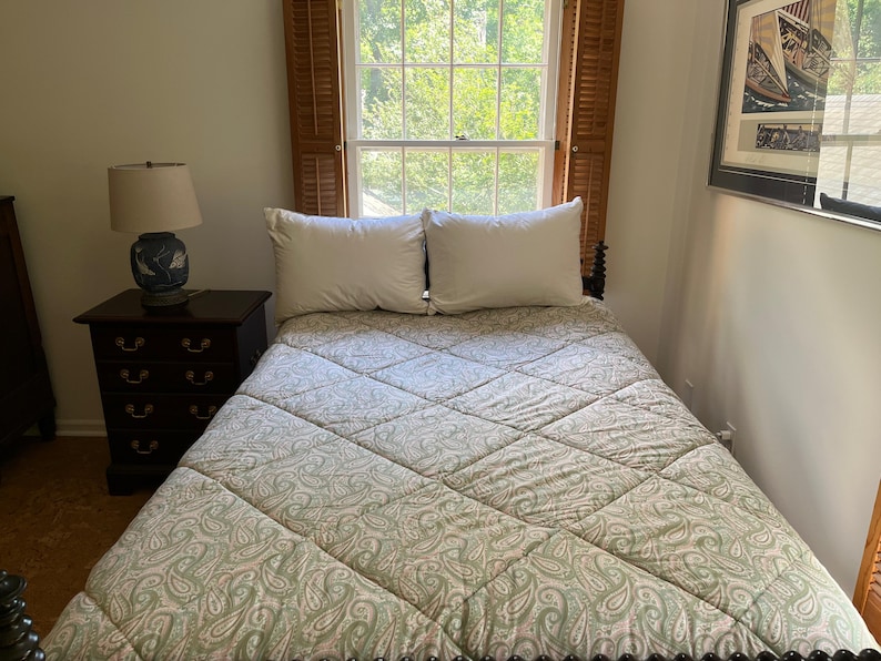 May include: A bed featuring a quilted comforter with a light green and white paisley pattern, two white pillows, and a dark wood nightstand with a blue and white ceramic lamp. The bed is positioned in front of a window with wooden shutters.