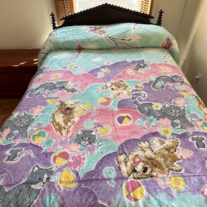 May include: A purple, pink, and teal quilted bedspread with a playful design featuring kittens and puppies playing with balls. The background features a cloud-like pattern.