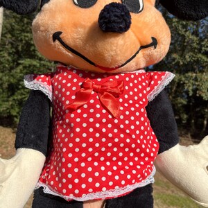Op de afbeelding: Een Minnie Mouse pluche pop met een perzikkleurig gezicht, zwarte oren en een zwarte neus. Ze draagt een rode polkadot jurk met een bijpassende strik en witte kanten rand. De pop heeft zwarte armen en benen en cr&egrave;mekleurige handen.