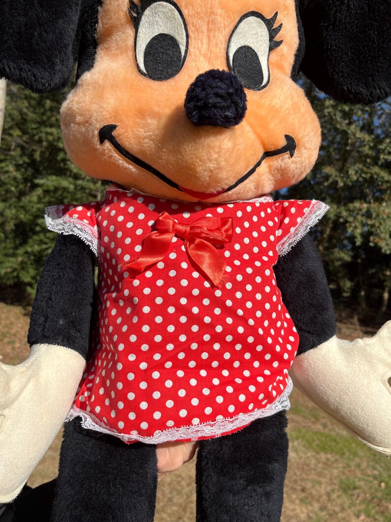Op de afbeelding: Een Minnie Mouse pluche pop met een perzikkleurig gezicht, zwarte oren en een zwarte neus. Ze draagt een rode polkadot jurk met een bijpassende strik en witte kanten rand. De pop heeft zwarte armen en benen en cr&egrave;mekleurige handen.