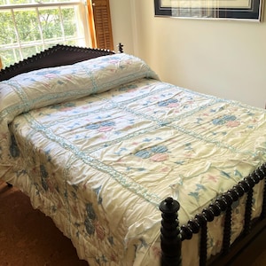 May include: A dark brown wooden bed frame with a spindle headboard and footboard, featuring a white quilt with a light blue ruffled grid pattern and floral motifs in shades of pink and blue, covering the mattress and pillows.