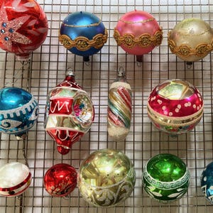 May include: A collection of vintage Christmas ornaments in various colors and designs. The ornaments include red, blue, pink, and gold spheres, some with glitter, stripes, and decorative trim. The ornaments are hanging on a metal grid.