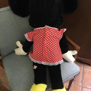 Op de afbeelding: Een pluche Minnie Mouse pop, van achteren gezien, zittend op een stoel. De pop heeft grote zwarte oren, een rode jurk met witte stippen en witte rand, zwarte benen, gele voeten en witte handschoenen. Een rode strik siert de bovenkant van het hoofd.