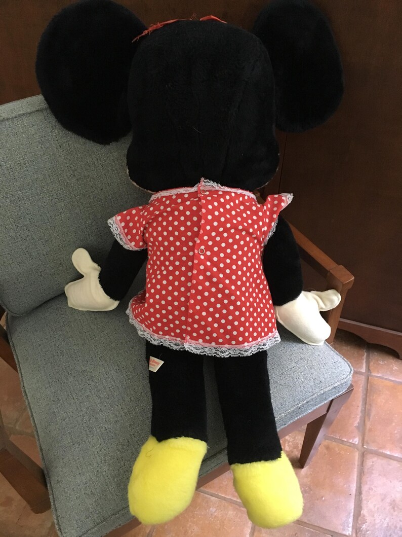 Op de afbeelding: Een pluche Minnie Mouse pop, van achteren gezien, zittend op een stoel. De pop heeft grote zwarte oren, een rode jurk met witte stippen en witte rand, zwarte benen, gele voeten en witte handschoenen. Een rode strik siert de bovenkant van het hoofd.