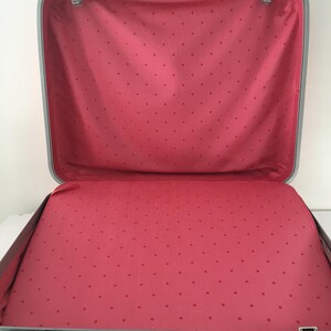 Pu&ograve; includere: Una valigia rigida aperta e vuota con una cornice color argento e una fodera interna rosa con un motivo a piccoli pois rosa scuro. La valigia &egrave; dotata di una maniglia rosa e di chiusure metalliche sui bordi superiore e inferiore.
