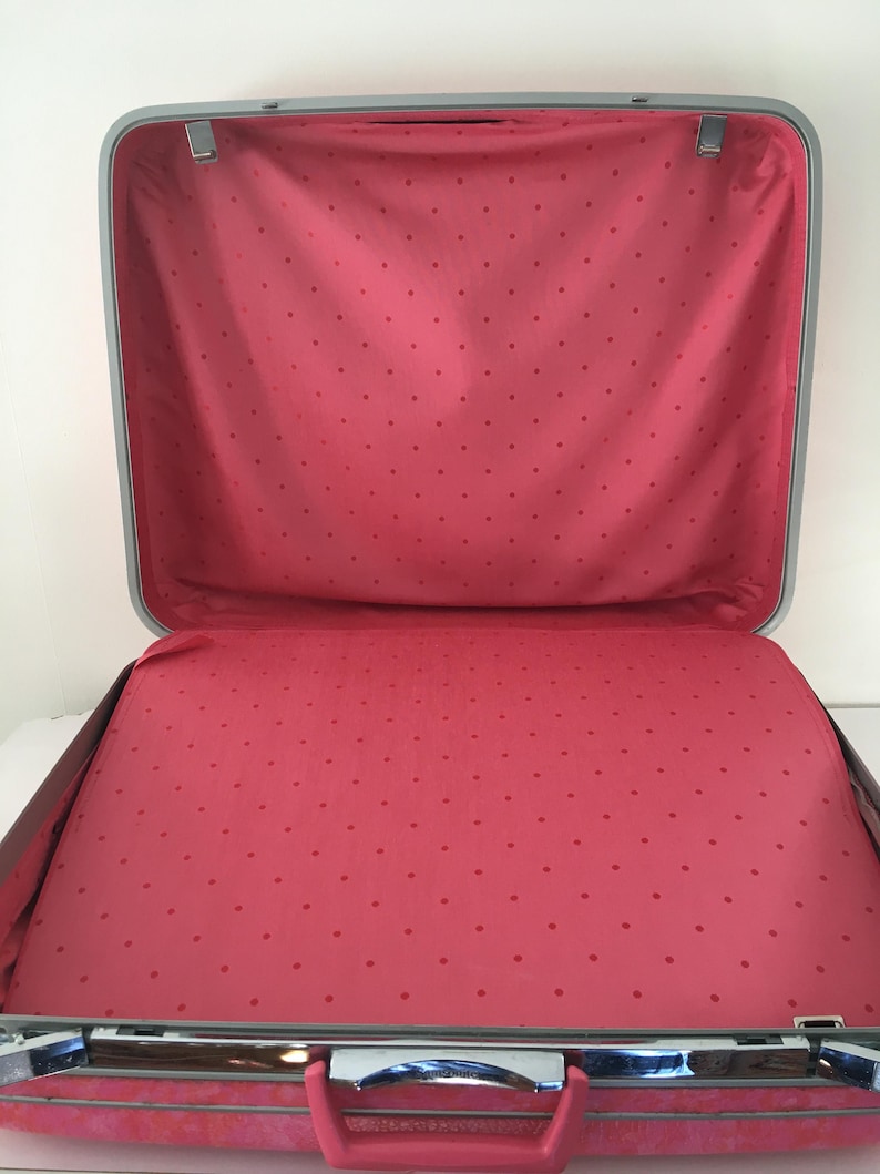 Pu&ograve; includere: Una valigia rigida aperta e vuota con una cornice color argento e una fodera interna rosa con un motivo a piccoli pois rosa scuro. La valigia &egrave; dotata di una maniglia rosa e di chiusure metalliche sui bordi superiore e inferiore.