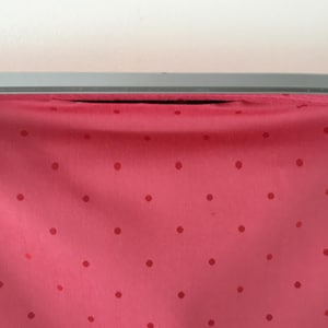 Pu&ograve; includere: Un tessuto rosa vivace caratterizzato da un motivo ripetitivo di piccoli pois rosso scuro. Il tessuto &egrave; drappeggiato e fissato a una cornice metallica grigia, liscia e orizzontale, che corre lungo il bordo superiore dell'immagine.