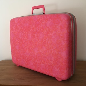 Pu&ograve; includere: Una valigia rigida rettangolare con un vivace motivo maculato rosa e arancione. Il bagaglio include una maniglia superiore rosa a tinta unita, una chiusura a zip color argento e piccoli piedini integrati sulla base.