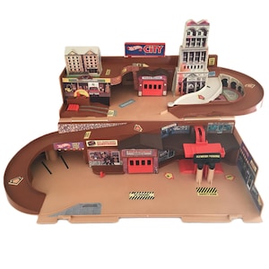 May include: A multi-level Hot Wheels City toy playset featuring brown plastic ramps, a police station, a tall building, and a red elevator parking mechanism. The structure includes various printed signs like Hot Wheels City, Hamburgers, and No Parking.