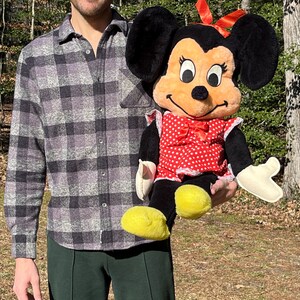 Op de afbeelding: Een pluche Minnie Mouse pop, met een rood-witte polkadot jurk, een rode strik en gele schoenen. De pop heeft zwarte oren en een lachend gezicht. De pop wordt vastgehouden tegen een natuurlijke buitenachtergrond.
