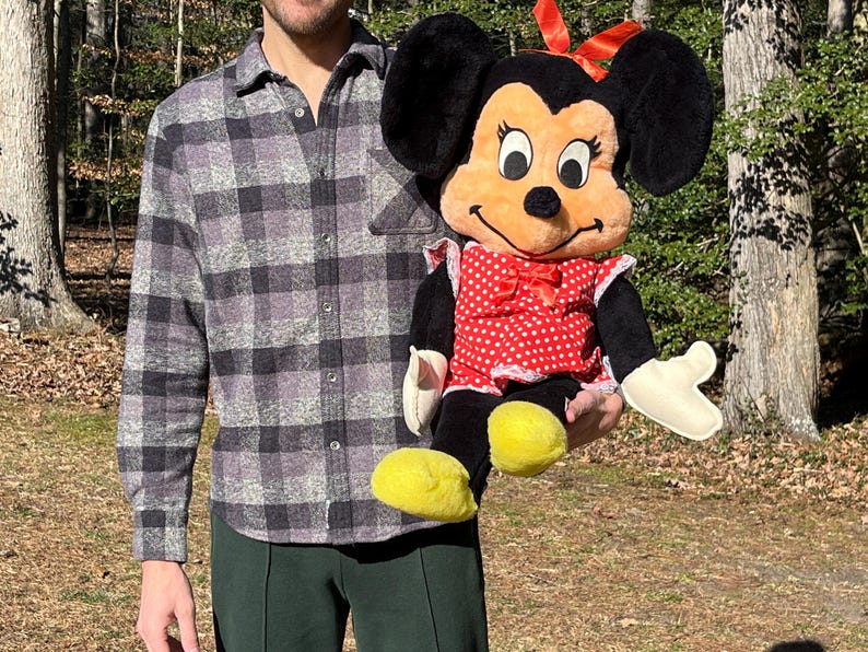 Op de afbeelding: Een pluche Minnie Mouse pop, met een rood-witte polkadot jurk, een rode strik en gele schoenen. De pop heeft zwarte oren en een lachend gezicht. De pop wordt vastgehouden tegen een natuurlijke buitenachtergrond.