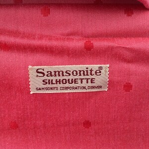 Pu&ograve; includere: Una fodera in tessuto rosa acceso con un sottile motivo a pois tono su tono. Al centro si trova un'etichetta rettangolare in tessuto beige con il testo Samsonite SILHOUETTE SAMSONITE CORPORATION, DENVER in un carattere serif marrone scuro.