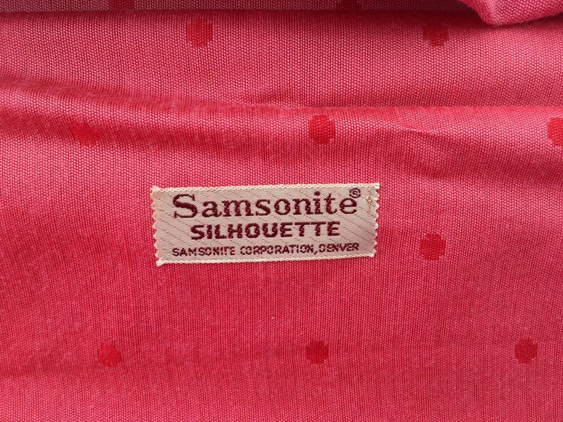 Pu&ograve; includere: Una fodera in tessuto rosa acceso con un sottile motivo a pois tono su tono. Al centro si trova un'etichetta rettangolare in tessuto beige con il testo Samsonite SILHOUETTE SAMSONITE CORPORATION, DENVER in un carattere serif marrone scuro.