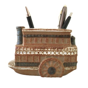 May include: A brown ceramic desk organizer shaped like a paddlewheel steamboat with a textured finish. The vessel features a prominent side wheel, a chimney, and horizontal detailing. Three pens are inserted into the top of the structure.