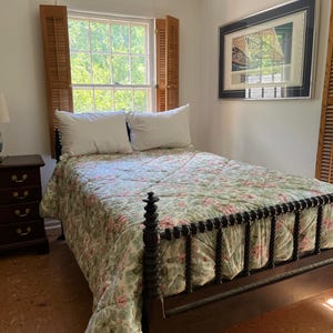 May include: A dark wood bed frame with turned spindle posts and a floral quilted bedspread in shades of green and pink. Two white pillows rest against the headboard. A dark wood nightstand with four drawers and a blue and white lamp sit nearby.