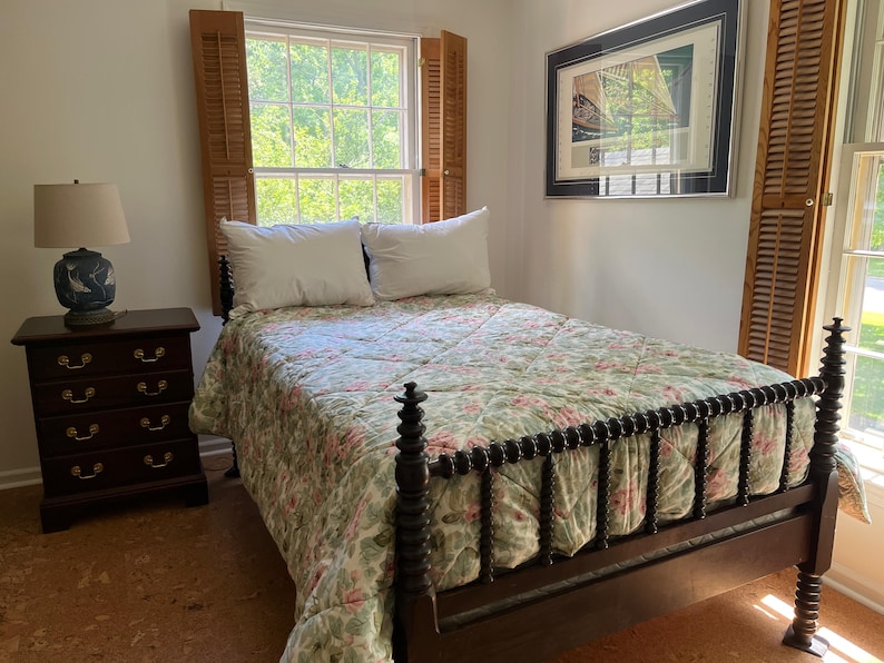 May include: A dark wood bed frame with turned spindle posts and a floral quilted bedspread in shades of green and pink. Two white pillows rest against the headboard. A dark wood nightstand with four drawers and a blue and white lamp sit nearby.