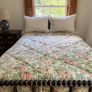 May include: A bed with a diamond-quilted comforter featuring a pink rose and green leaf floral pattern, two white pillows, and a dark wood spindle headboard. Beside the bed sits a dark wood nightstand with a blue and white ceramic table lamp.