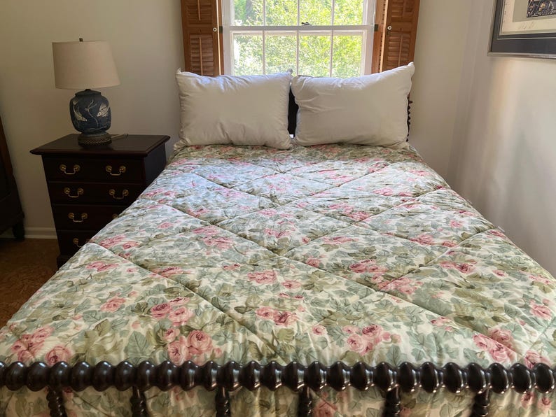 May include: A bed with a diamond-quilted comforter featuring a pink rose and green leaf floral pattern, two white pillows, and a dark wood spindle headboard. Beside the bed sits a dark wood nightstand with a blue and white ceramic table lamp.