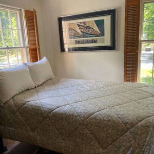 May include: A bed with a dark brown wooden spindle frame, topped with a light beige quilted comforter featuring a subtle paisley pattern and two white pillows. A framed print of sailboats hangs on the wall above the headboard between two windows.