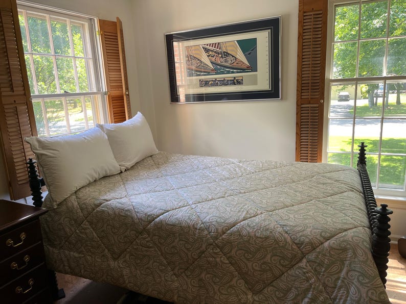 May include: A bed with a dark brown wooden spindle frame, topped with a light beige quilted comforter featuring a subtle paisley pattern and two white pillows. A framed print of sailboats hangs on the wall above the headboard between two windows.
