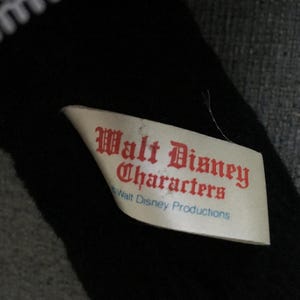 Op de afbeelding: Een zwart pluche speelgoed met een witte kanten rand. Een wit label met rode tekst luidt "Walt Disney Characters" en "Walt Disney Productions". Het speelgoed ligt op een grijze ondergrond.