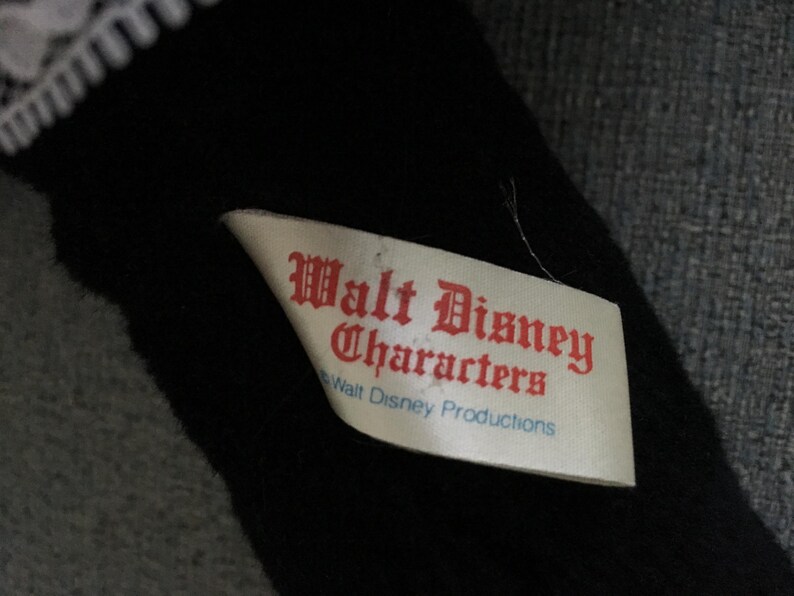 Op de afbeelding: Een zwart pluche speelgoed met een witte kanten rand. Een wit label met rode tekst luidt "Walt Disney Characters" en "Walt Disney Productions". Het speelgoed ligt op een grijze ondergrond.