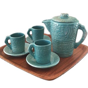 May include: A turquoise ceramic tea set with a matching teapot, three cups, and saucers. The set features embossed designs and is displayed on a wooden tray. The teapot has a lid and a handle.