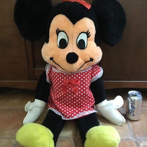 Op de afbeelding: Een vintage Minnie Mouse pluche pop met zwarte oren, een rode strik en een rood-witte polkadot jurk. De pop heeft witte handschoenen en gele voeten. Een Pepsi-blikje staat naast de pop.