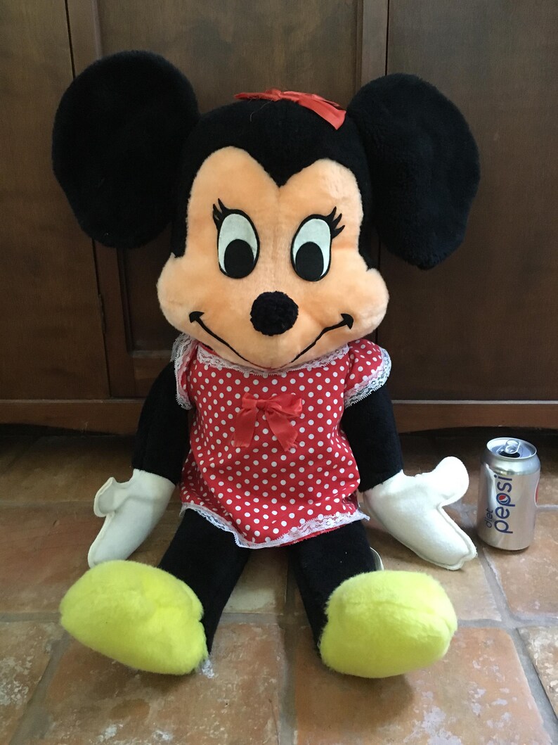 Op de afbeelding: Een vintage Minnie Mouse pluche pop met zwarte oren, een rode strik en een rood-witte polkadot jurk. De pop heeft witte handschoenen en gele voeten. Een Pepsi-blikje staat naast de pop.