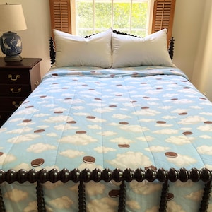 May include: A bed featuring a light blue quilt patterned with white clouds and brown circular sandwich cookies. The bed has two white pillows and a dark brown wooden spindle headboard and footboard, with a matching dark brown nightstand nearby.