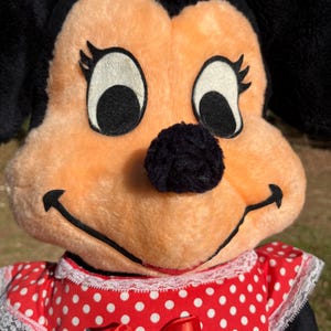Op de afbeelding: Close-up van een Minnie Mouse pluche pop. De pop heeft een perzikkleurig gezicht, zwarte oren en een zwarte neus. Ze draagt een rode jurk met witte stippen en een rode strik. De pop heeft grote, zwarte ogen en een glimlachende uitdrukking.